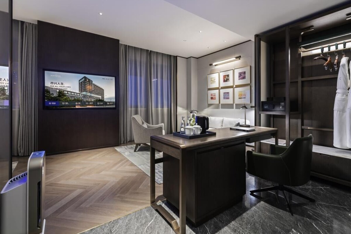 A modern hotel room with a flat-screen TV, a desk with a chair, a wardrobe, and a large window with curtains.