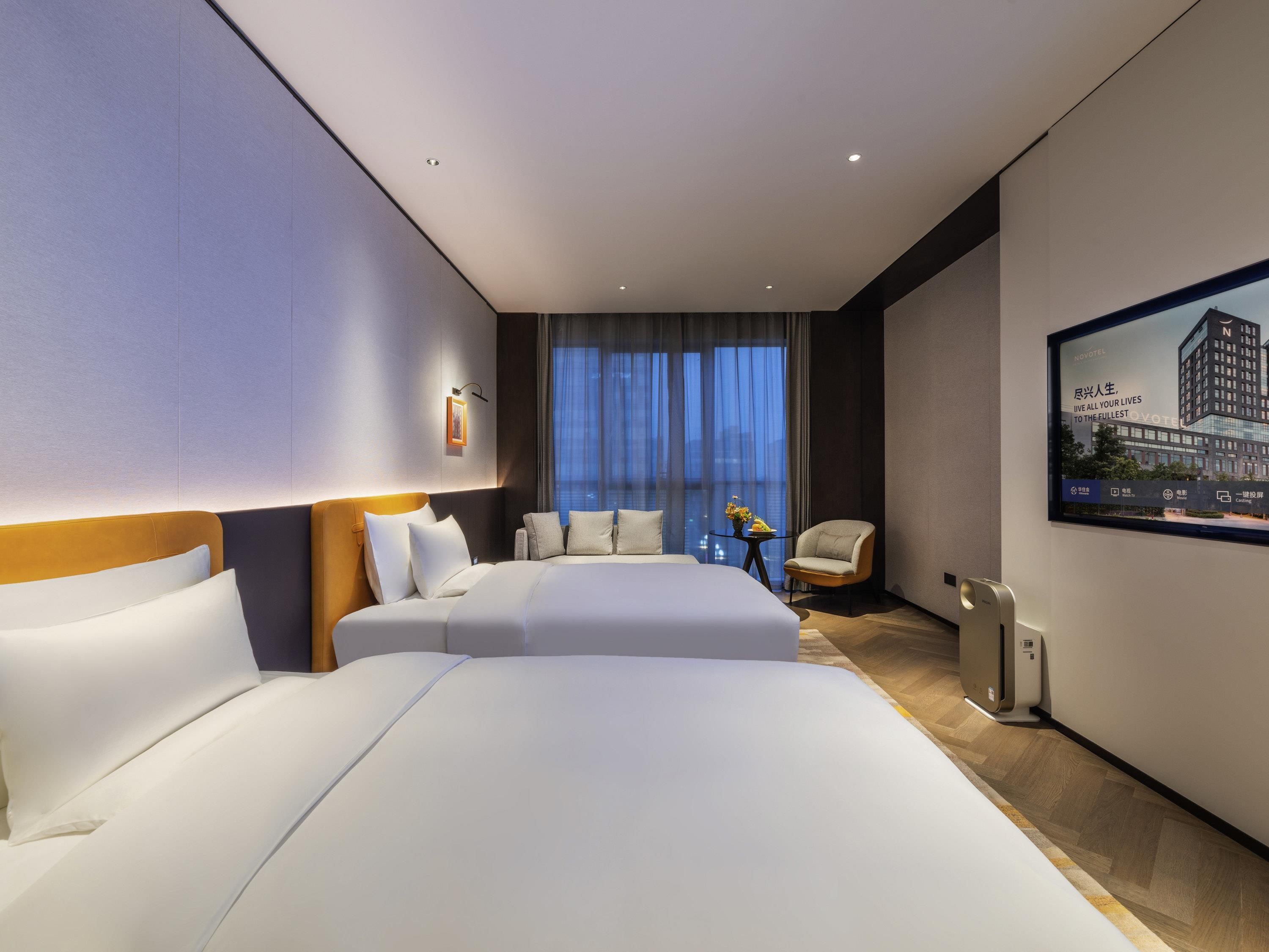 A modern hotel room with two beds, a seating area, a view of the city, and a large window.