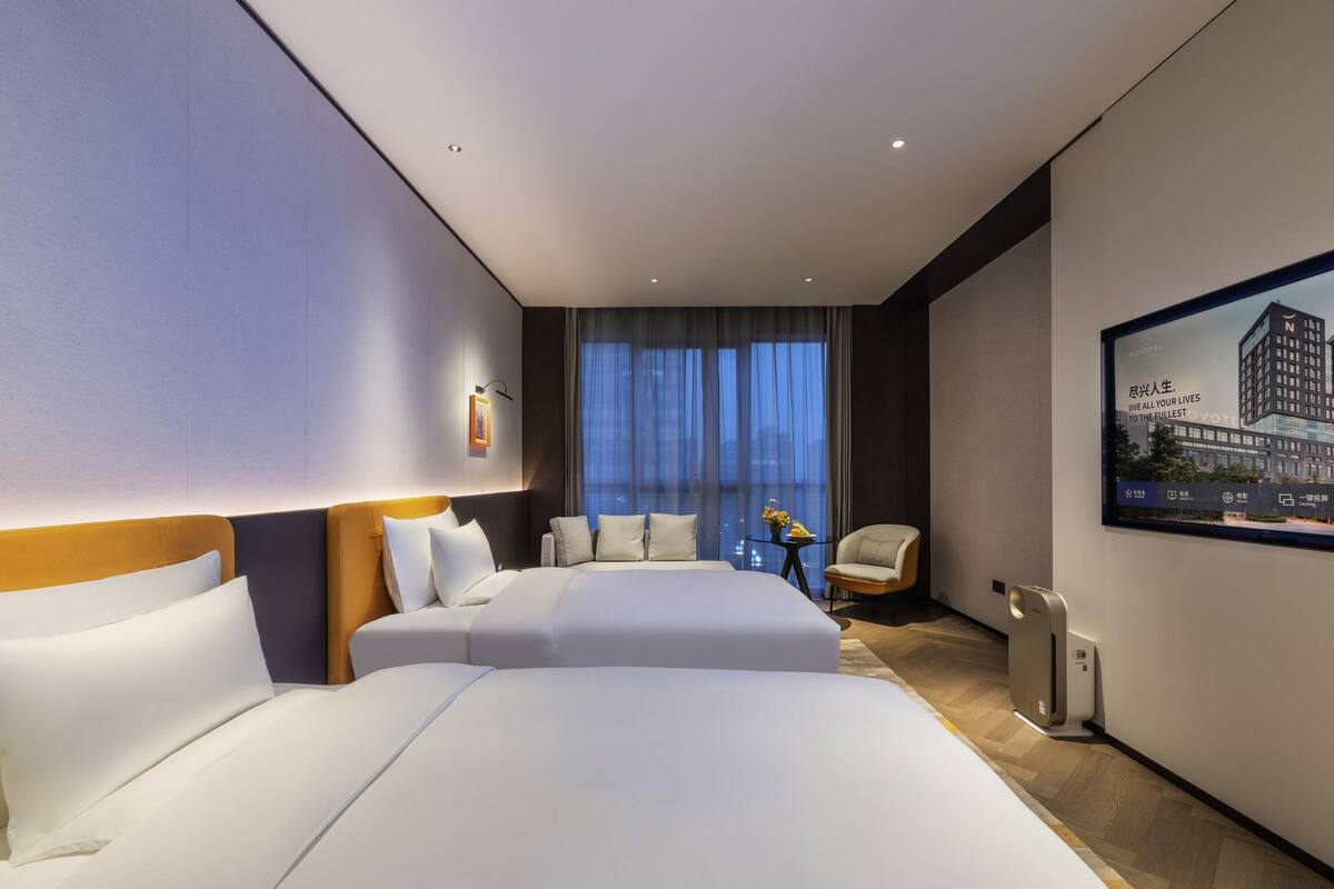 A modern hotel room with two beds, a seating area, a view of the city, and a large window.