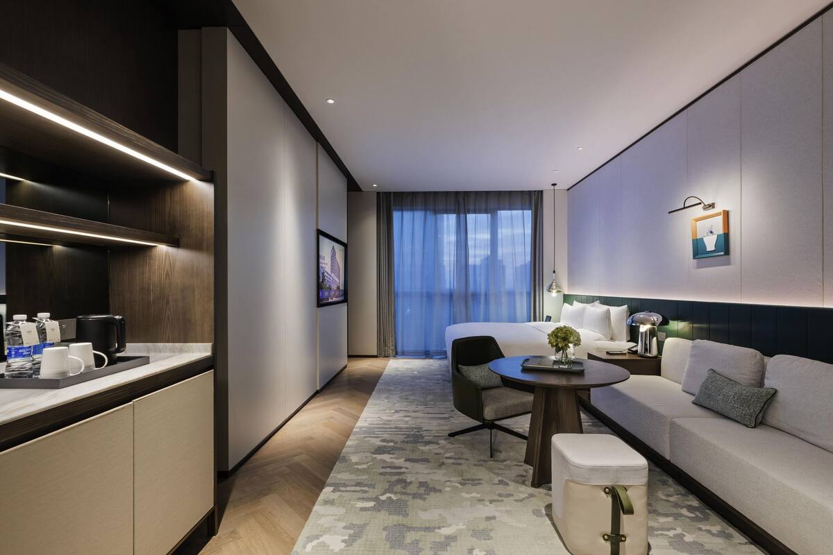 A modern hotel room with a bed, a sofa, a dining table, a kitchen area, and a large window with curtains.