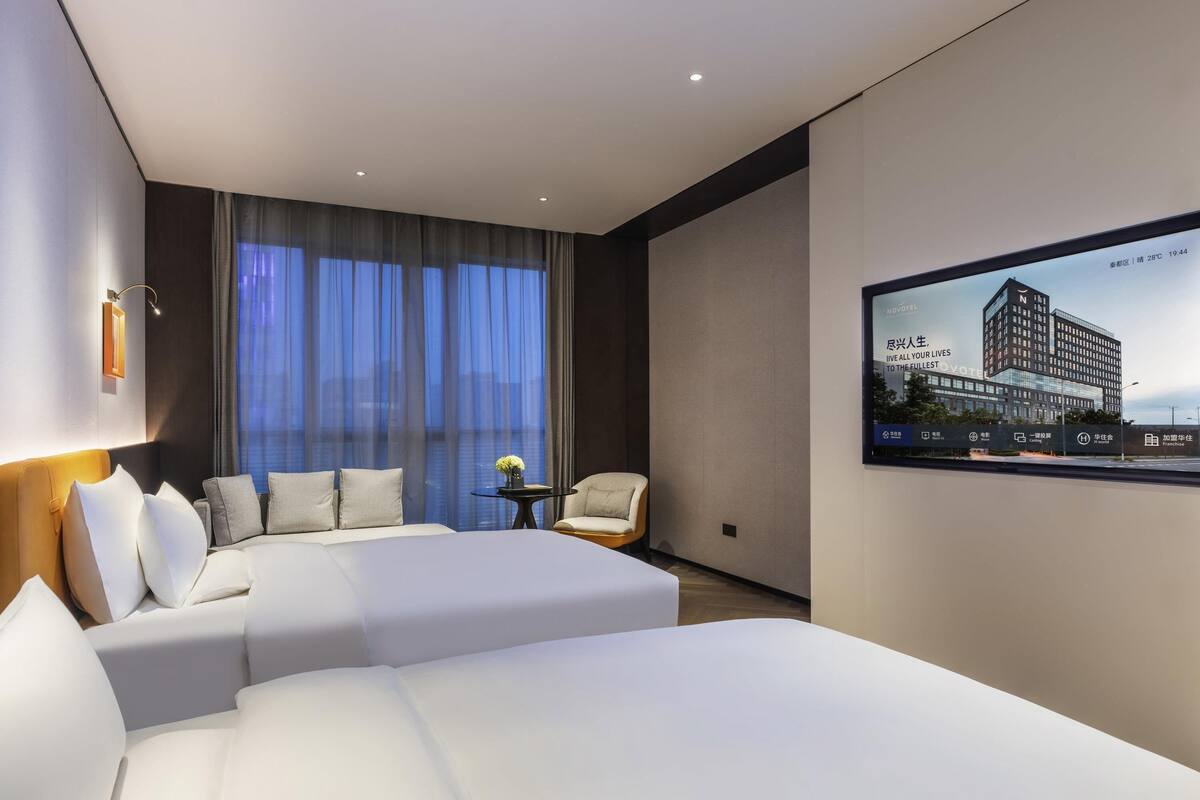 A modern hotel room with a large bed, a sofa, a small table with a vase, and a large window with curtains.