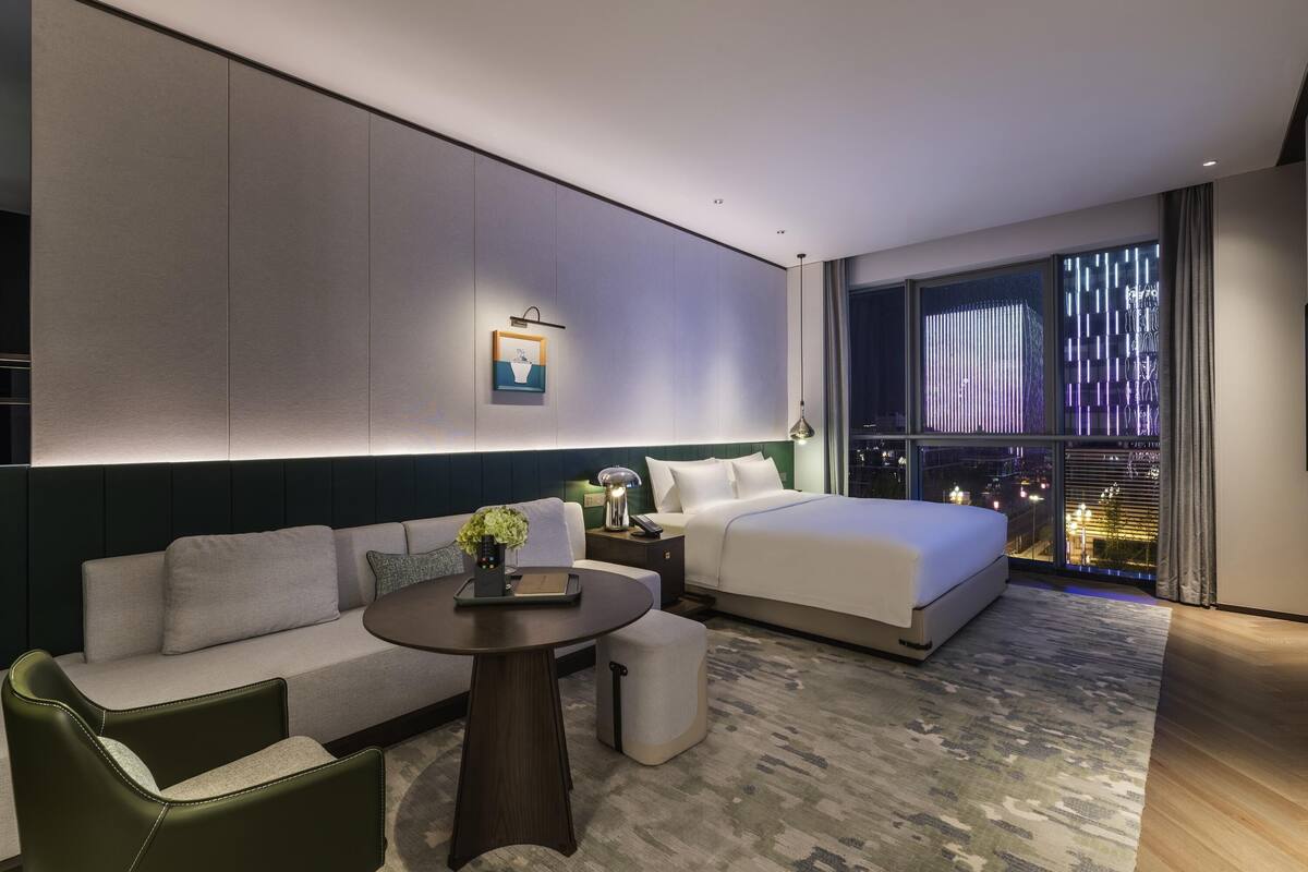 A modern hotel room with a bed, a sofa, a small table, and a view of the city at night.