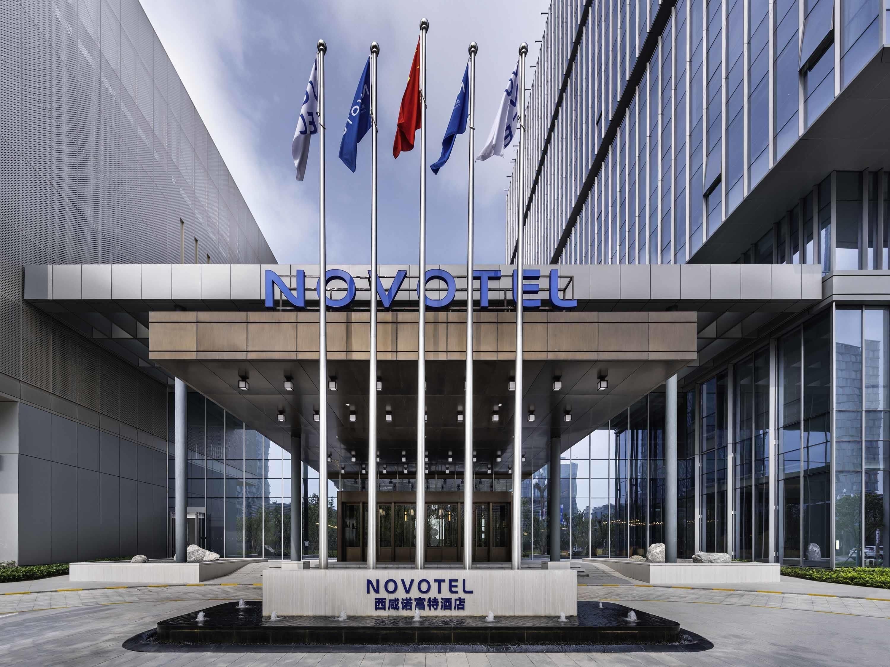 Une entrée d’hôtel moderne, avec l’enseigne « NOVOTEL », plusieurs drapeaux et une fontaine à l’avant.