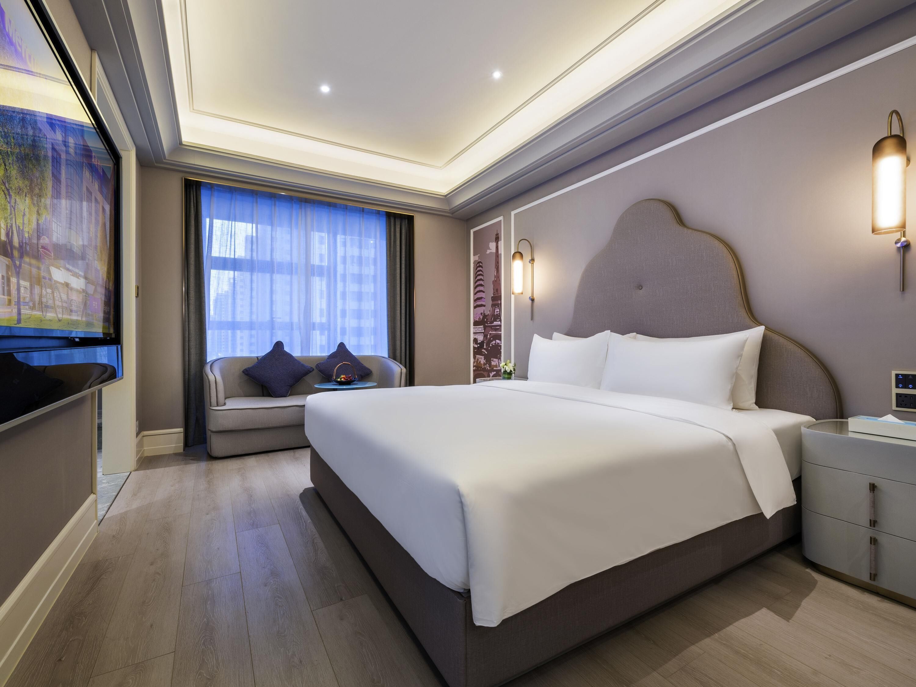 A modern hotel room with a large bed, a sofa with cushions, a TV, and a window with curtains.