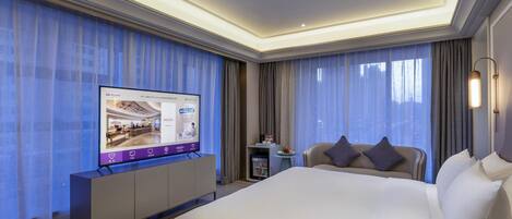 A hotel room with a large bed, a TV on a cabinet, a small table with fruit, and a sofa with cushions.