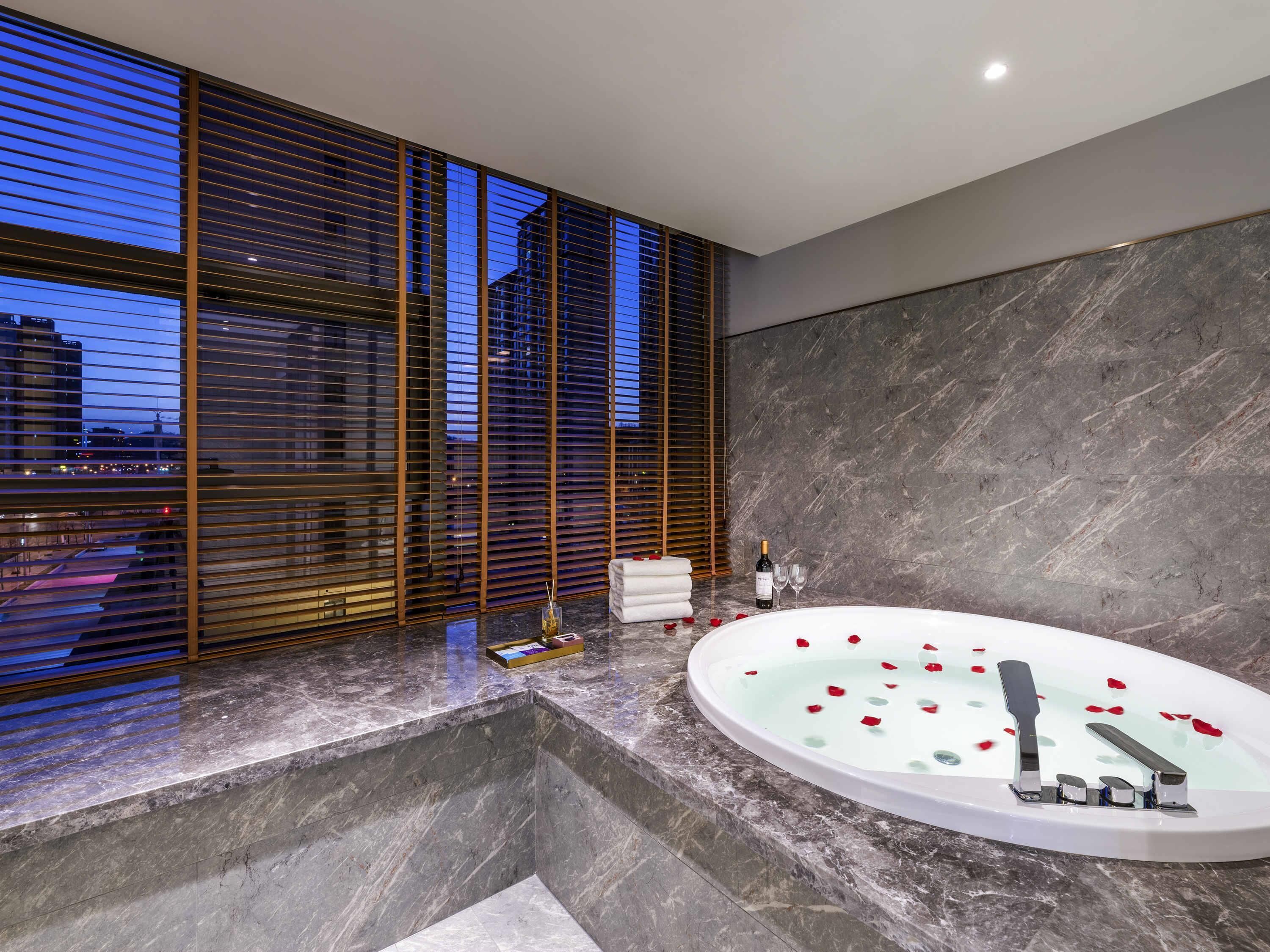 A modern bathroom with a large bathtub, marble countertops, and a city view through floor-to-ceiling windows with blinds.