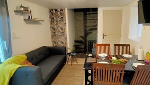 A living room with a sofa, a dining table set for four, and a staircase leading to an upper floor. - Maison au Calme à Proximité des Plages (Querrien)