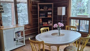 Ein Holzblockhaus-Interieur mit einem runden Tisch, gelben Stühlen, einem Bücherregal und einem Fenster mit Blick auf verschneite Bäume.