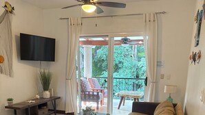 A living room with a sofa, armchair, coffee table, TV, and a balcony with a view of greenery. - Jazmin Del Sol - Just Steps Away to the Beautiful Potrero Beach (Potrero)