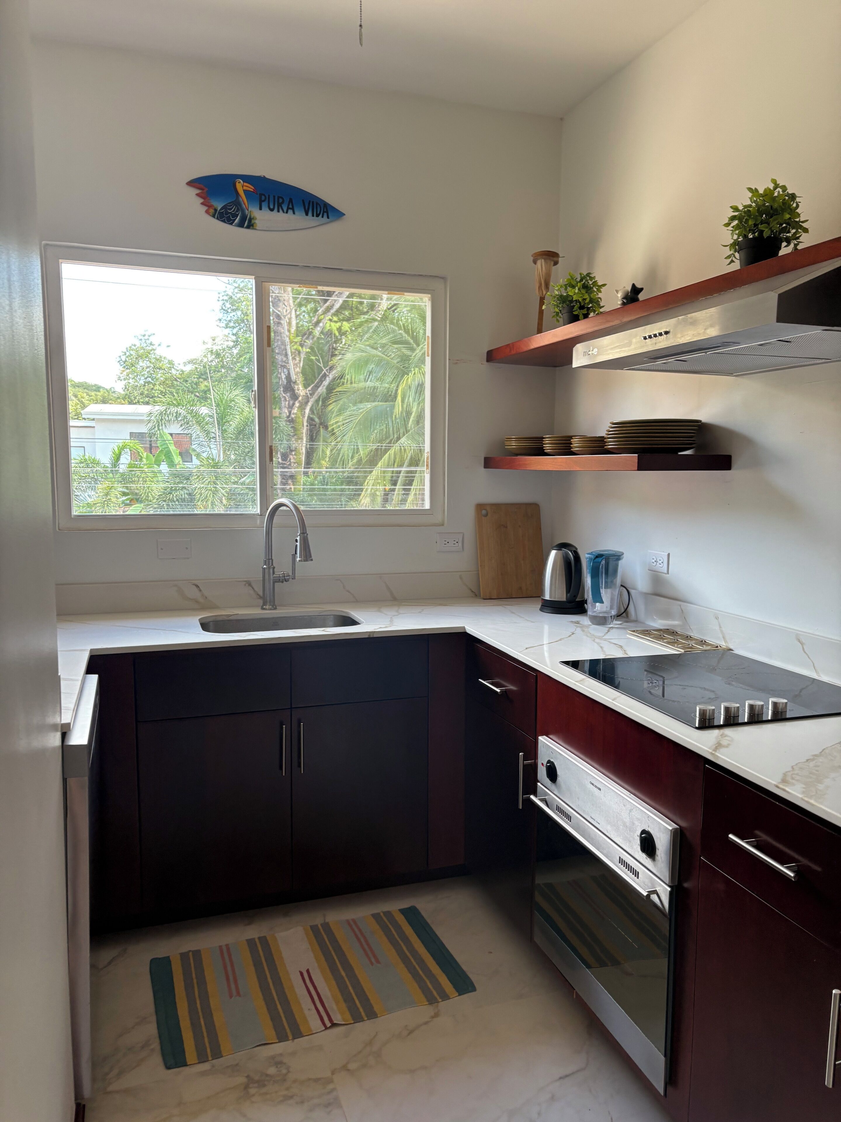 A compact kitchen with a sink, stove, and oven, a window with a view of greenery, and a decorative sign above the window.
