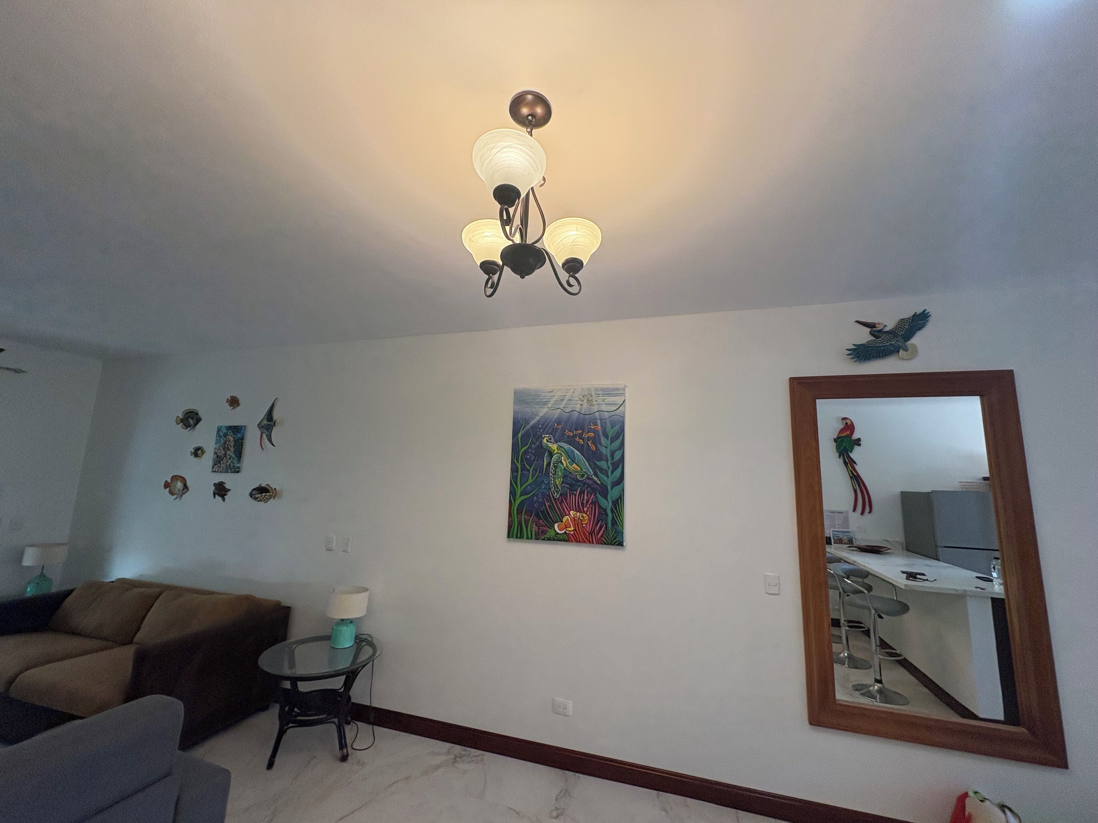 Een woonkamer met een bank, een klein rond tafeltje met een lamp, een schilderij van een onderwaterlandschap en een grote spiegel die een keuken weerkaatst.