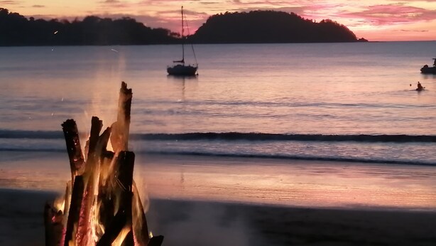 A calm beach scene with a wooden structure in the foreground, a sailboat in the distance, and a hilly landscape under a sunset sky. - Jazmin Del Sol - Just Steps Away to the Beautiful Potrero Beach (Potrero)