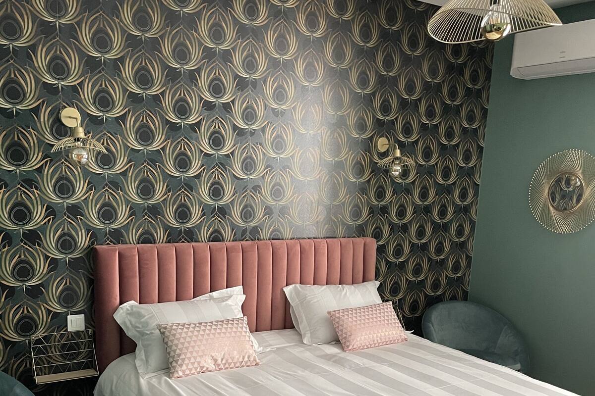 Een slaapkamer met behang met een patroon, een roze gestoffeerd hoofdeinde, wit beddengoed en een nachtkastje met een lamp.
