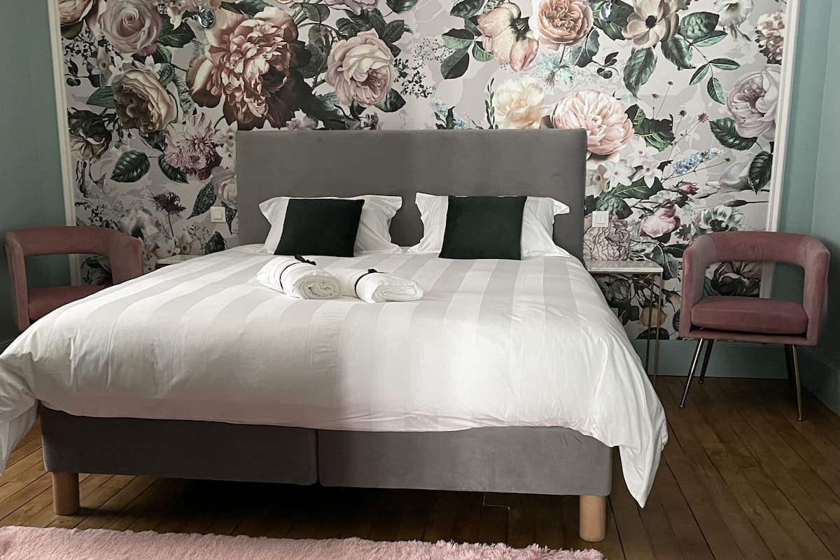 Een slaapkamer met een groot bed, bloemenbehang, twee stoelen en een roze vloerkleed.