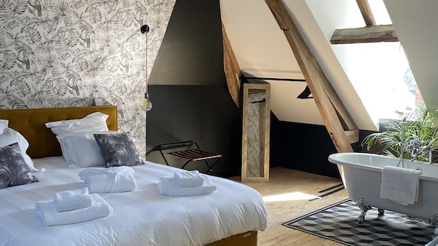 Une chambre avec un grand lit, un plafond en bois, un papier peint à motifs floraux en noir et blanc, une baignoire blanche et un tapis jaune.