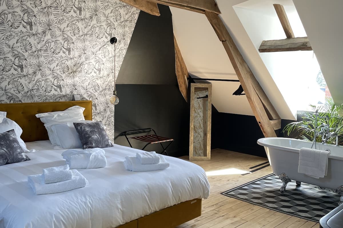 Een slaapkamer met een groot bed, een houten plafond, behangpapier met bloemen in zwart-wit, een badkuip in het wit en een geel vloerkleed.