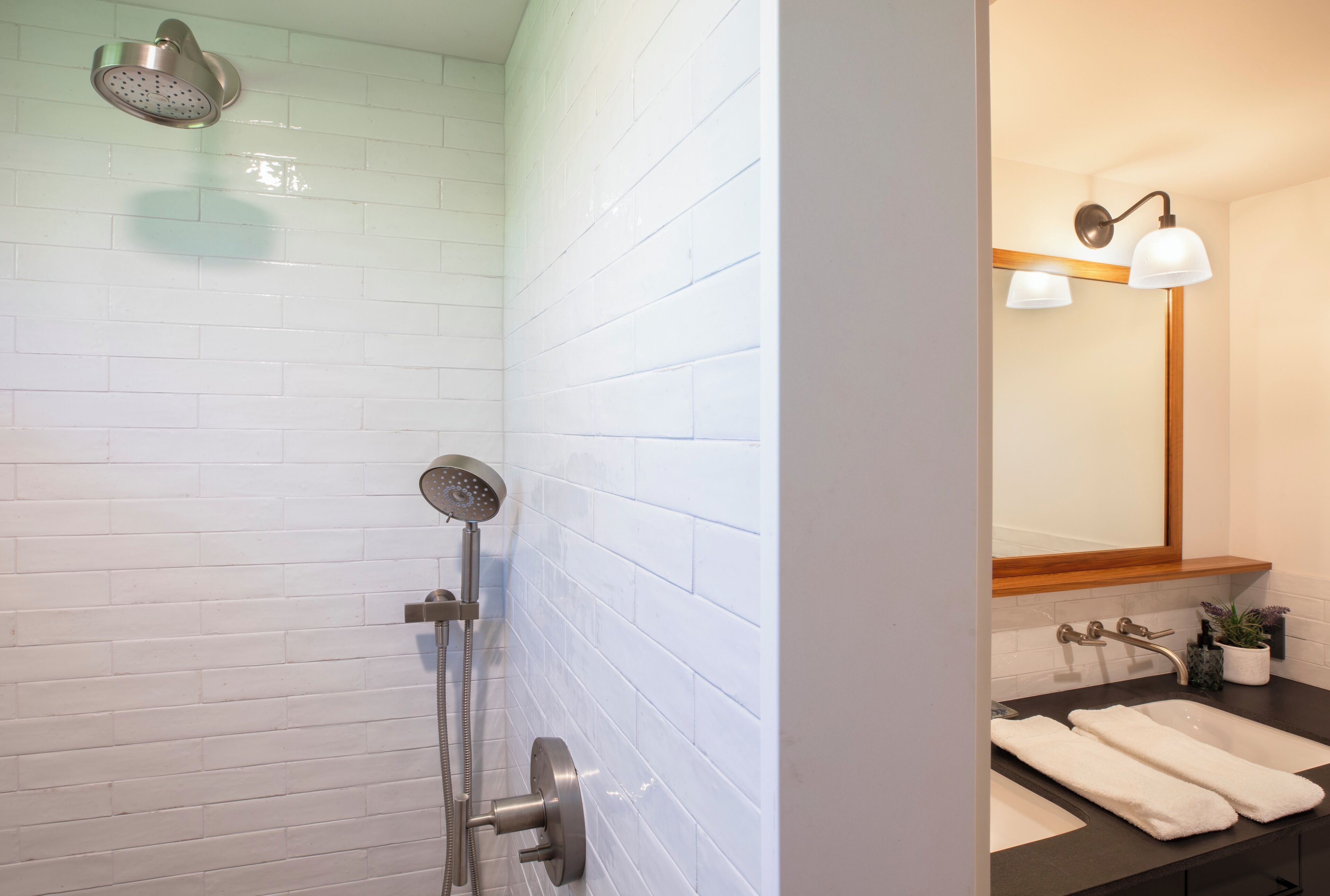 Une salle de bain avec des murs recouverts de carreaux blancs, une douche avec un pommeau de douche amovible, un lavabo avec un miroir et deux serviettes posées sur le comptoir.