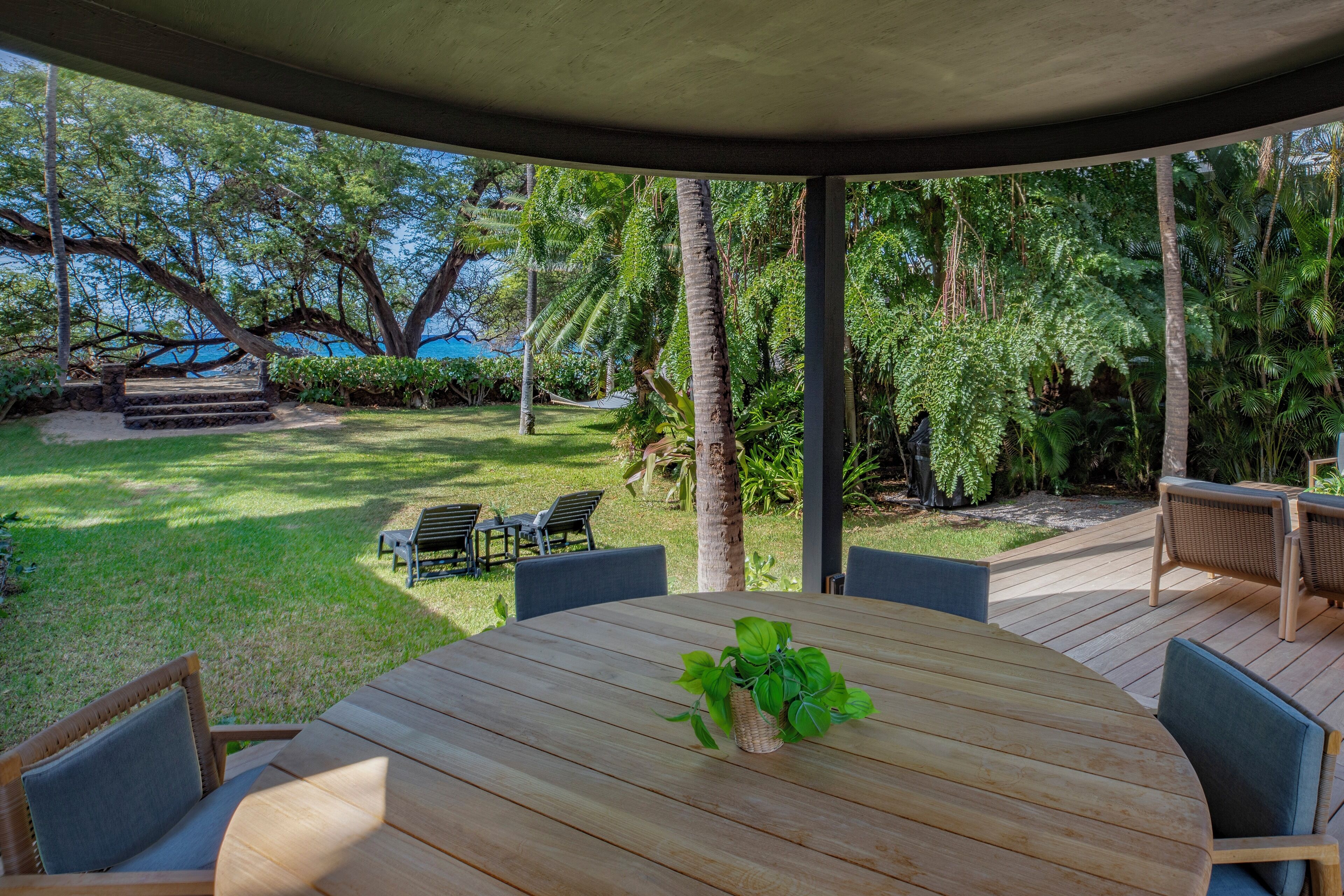 A covered outdoor dining area with a wooden table and chairs, surrounded by lush greenery and a view of the ocean.