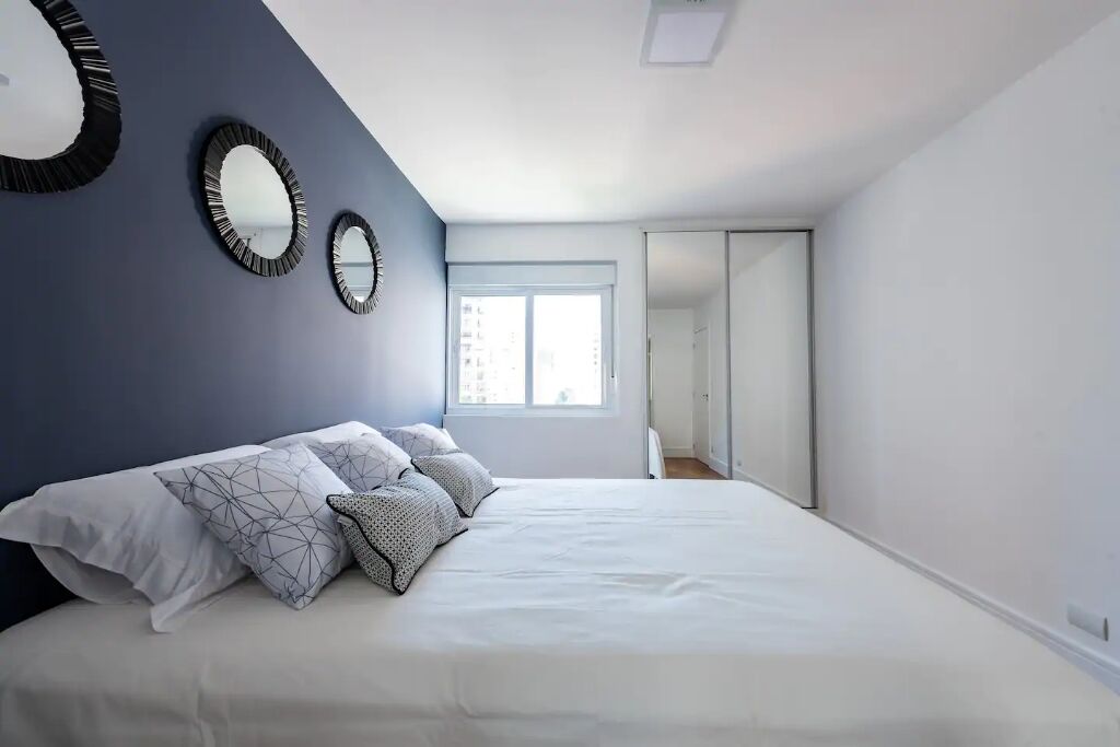 A bedroom with a bed, two wall mirrors, a window, and a door leading to another room.