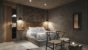 A modern bedroom with a brick accent wall, a bed with white linens, two wooden chairs, a small table, and a nightstand.