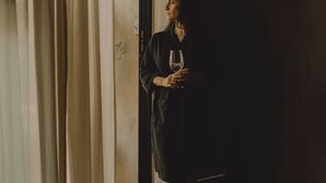 A person standing in a dimly lit room with a door, curtains, and a concrete floor.