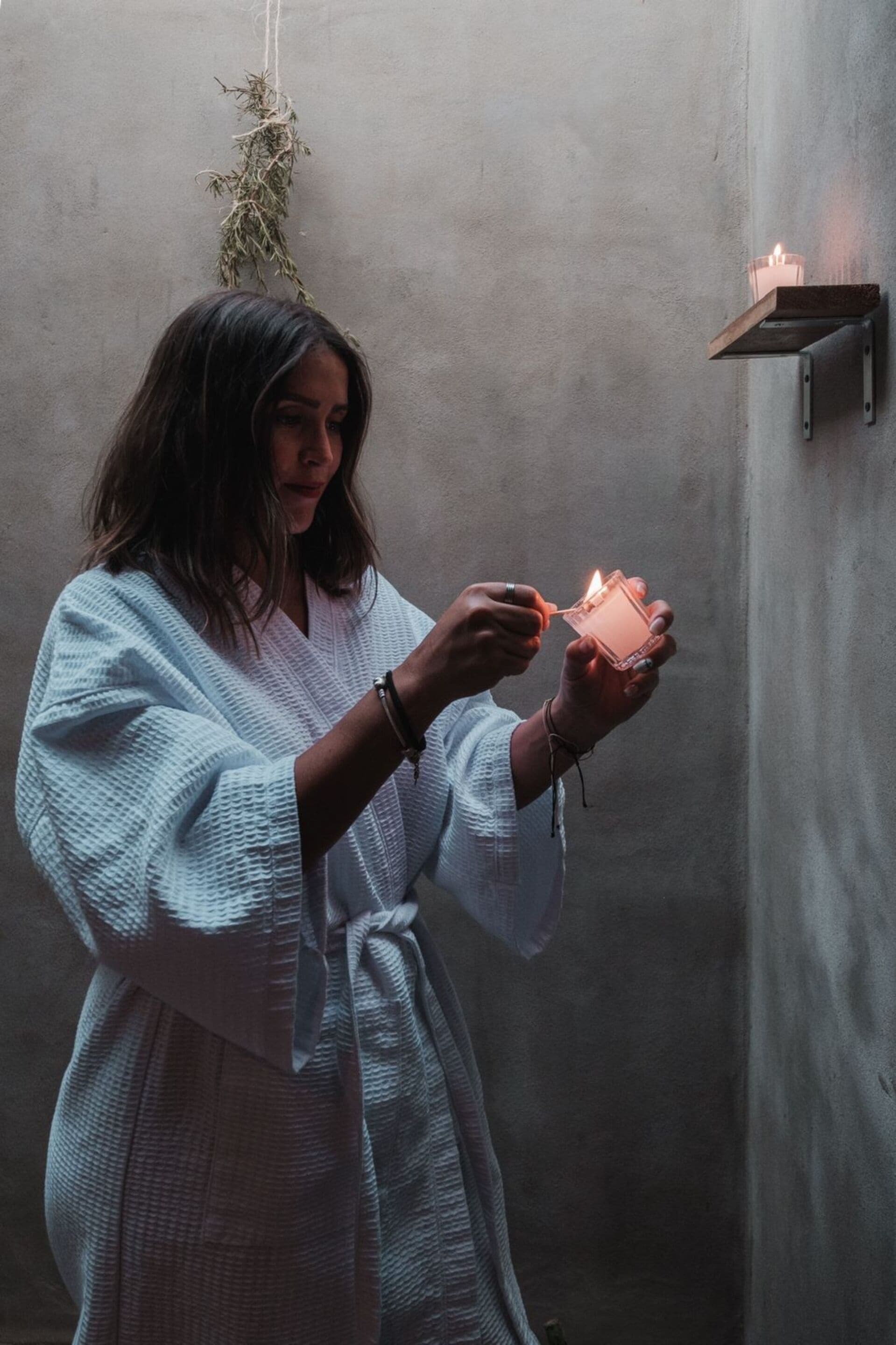 A person in a white robe lighting a candle in a dimly lit room with a concrete wall and a small shelf.