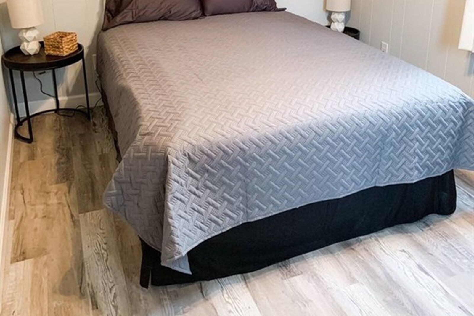 Un lit recouvert d’une couette grise, une petite table de chevet avec une lampe et un plancher en bois.