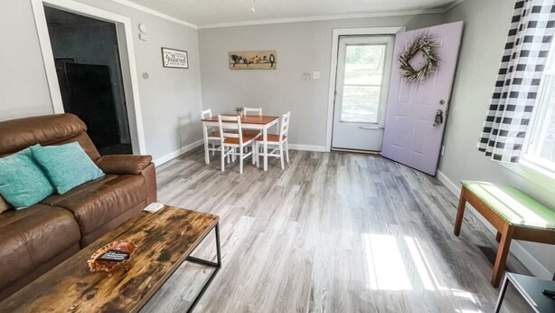 A living room with a brown leather sofa, a wooden coffee table, a dining table with chairs, a ceiling fan, and a door with a wreath.