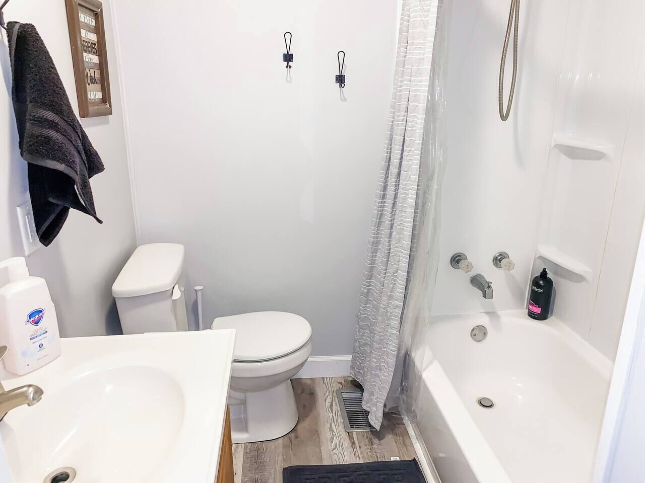 Un baño con inodoro, lavamanos y bañera. Hay dos sujetapapeles negros en la pared, una cortina de ducha y una botella de champú.