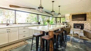 Une cuisine moderne dotée d’un grand îlot central, de tabourets de bar, d’une cheminée en pierre et d’une grande fenêtre donnant sur un espace verdoyant.
