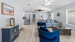 A spacious living room with a grey sofa, blue armchairs, and a wooden coffee table. A ceiling fan and a chandelier are visible.