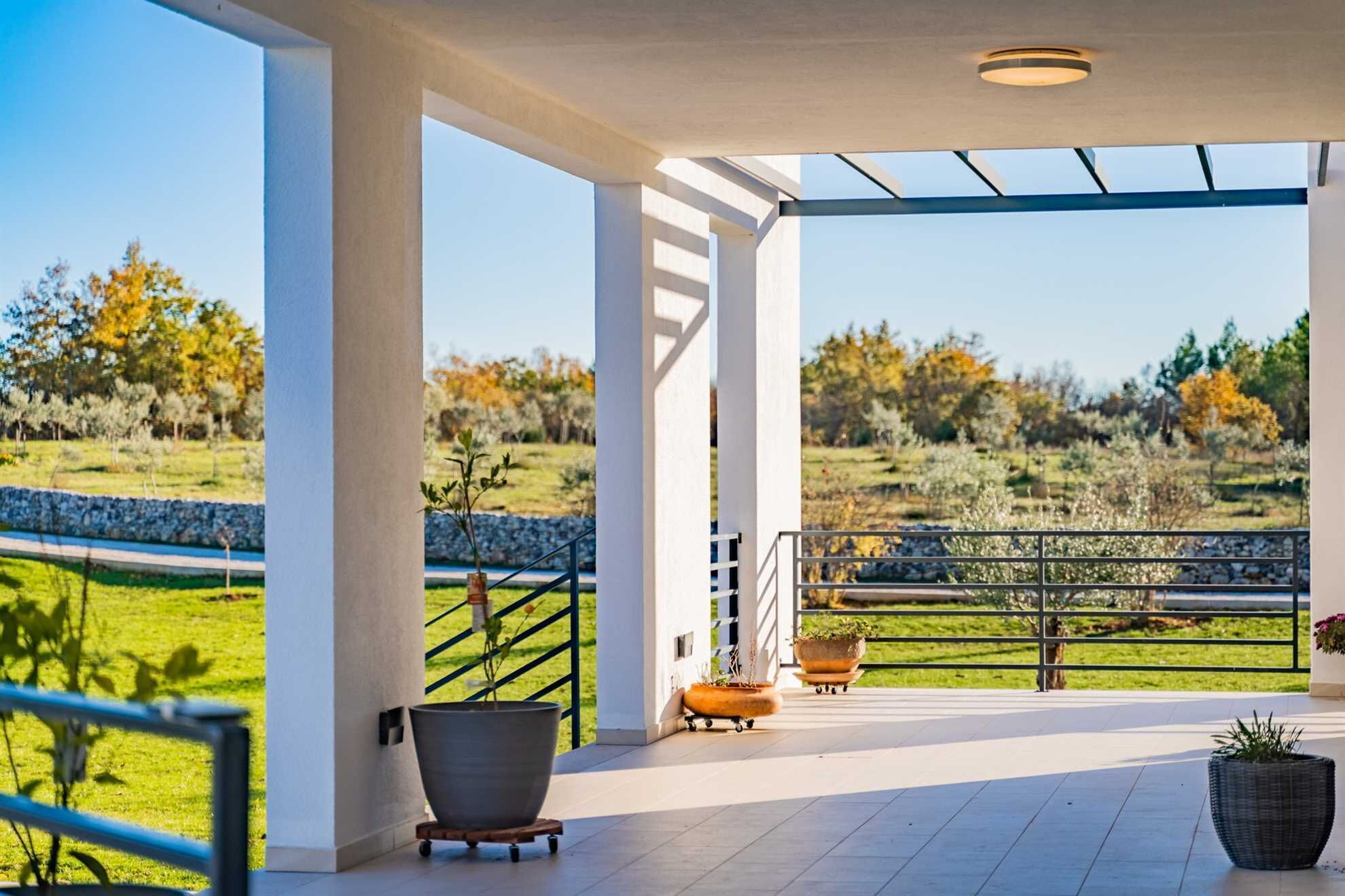 Un espace extérieur abrité, offrant une vue sur un champ verdoyant et des arbres. On y trouve des rampes en métal, un escalier et plusieurs jardinières.