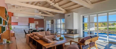 A spacious living area with a wooden ceiling, a fireplace, and large windows offering a view of the outdoors.