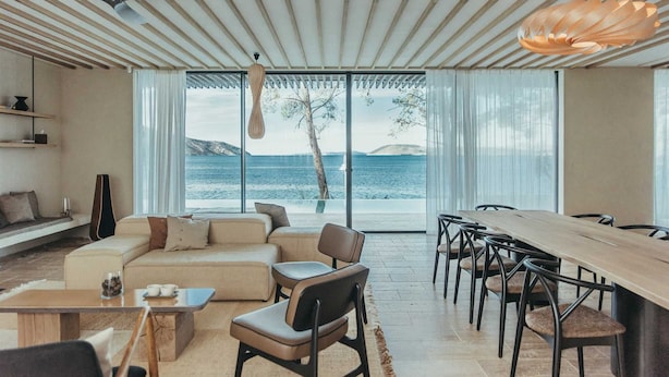 Un salon moderne doté d’une grande fenêtre donnant sur un plan d’eau, un coin repas avec une longue table et des chaises, et un espace salon confortable avec un canapé et des fauteuils.