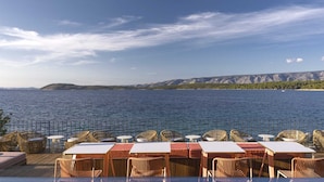 Un espace de restauration en plein air, avec des tables et des chaises en osier, offrant une vue sur une étendue d’eau et sur des montagnes lointaines.