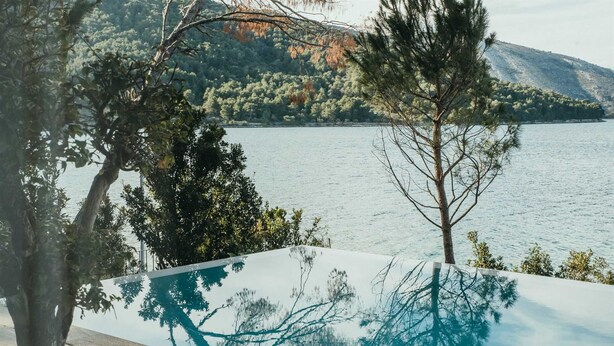 Vista incantevole su un lago, con un pontile di legno e una piscina a sfioro in vetro che riflette alberi e montagne.