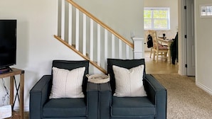 A living room with a dark green sofa set, a television on a wooden stand, a staircase with wooden railings, and a dining area in the background.
