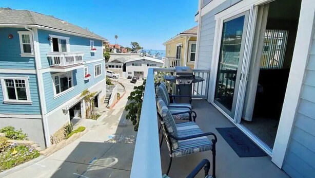 A residential balcony with a seating area, a grill, and a view of the street and neighboring houses.