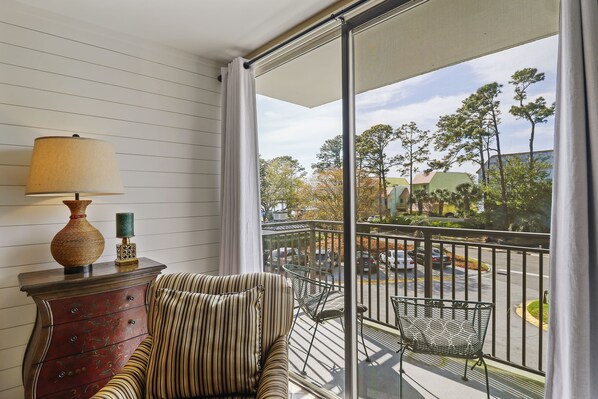 A balcony with a striped chair, a wooden cabinet with a lamp, and a view of a street and trees.