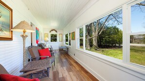 Une pièce longue et étroite, dotée d’un plancher en bois et de murs blancs, comprenant un canapé, des fauteuils, une table et de grandes fenêtres donnant sur l’extérieur.