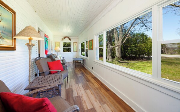 A long, narrow room with wooden flooring and white walls, featuring a sofa, chairs, a table, and large windows offering a view of the outdoors.