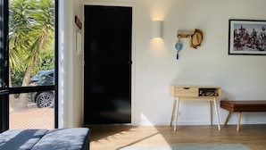 Ein Zimmer mit einer schwarzen Tür, einem kleinen Holztisch mit Schubladen, einer Bank, einer Wandleuchte und einem gerahmten Bild.