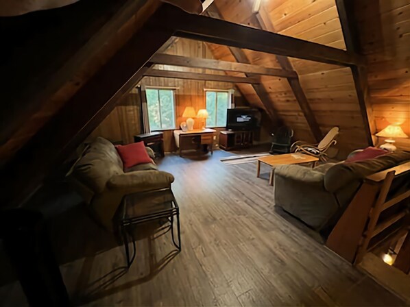 Relax in this charming attic room with a sofa, chair, and TV, set amidst a warm and inviting atmosphere.