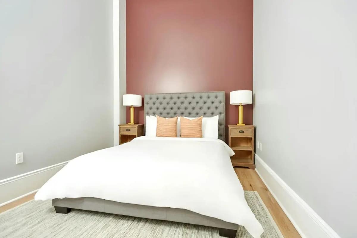 A neatly made bed with a grey headboard, two bedside tables with lamps, and a plain wall in the background.