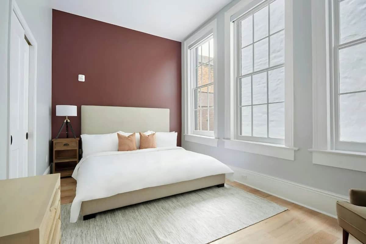 A bedroom with a large bed, a nightstand, a lamp, and a window with white frames.