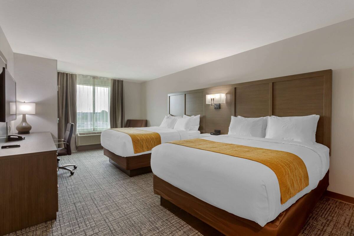 A hotel room with two beds, a desk, a chair, a lamp, and a window with curtains.