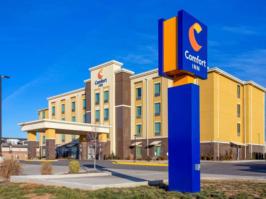 Un hotel Comfort Inn con fachada amarilla, un cartel azul llamativo y un estacionamiento con algunos arbustos pequeños.