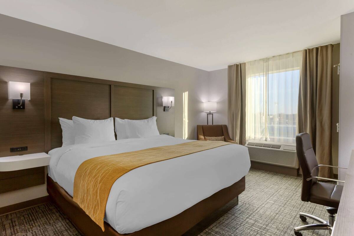 A hotel room with a large bed, bedside tables, a desk with a chair, a window with curtains, and wall-mounted lamps.