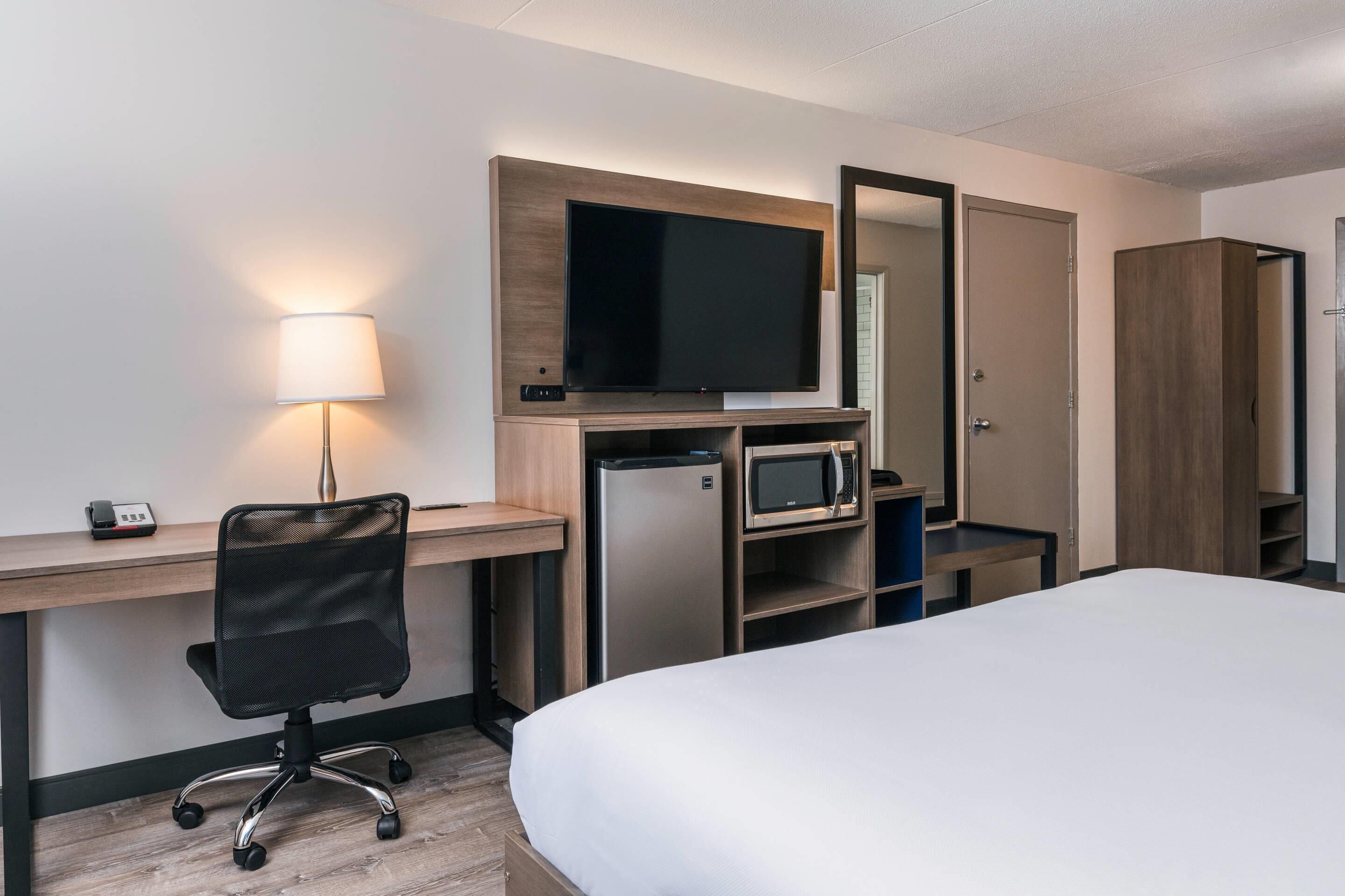 Quarto de hotel com cama, escrivaninha com computador, televisão, micro-ondas e frigorífico.