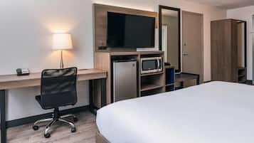 Quarto de hotel com cama, escrivaninha com computador, televisão, micro-ondas e frigorífico.